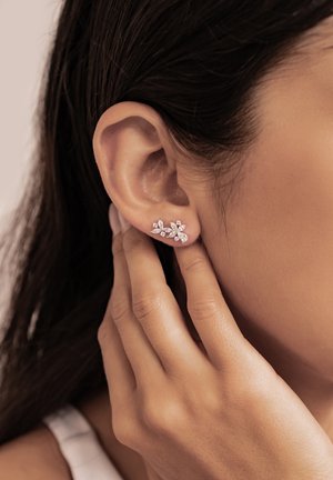 Person berührt Ohr, das mit zarten, blumenförmigen Diamantohrringen geschmückt ist, mit dunklem Haar und hellem Hautton sichtbar.