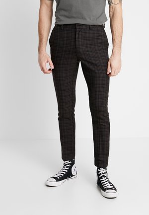 Hombre vistiendo pantalones ajustados a cuadros negros y marrones con zapatillas Converse altas negras sobre un fondo blanco.