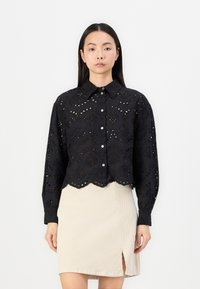Black cropped shirt with embroidering and eyelet cutouts, featuring a pointed collar, button closure, and scalloped hem. Paired with a beige skirt.