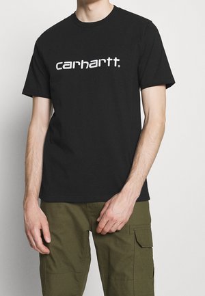 Personne portant un t-shirt noir Carhartt et un pantalon cargo vert olive, debout devant un fond clair uni.