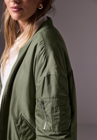 Oliivinvihreä bomber-takki, jossa on vetoketjullinen tasku hihassa, tikattu pintakuvio, resorikaulus ja väljä istuvuus, yhdistettynä valkoiseen paitaan.