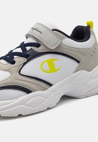 Scarpe sportive caratterizzate da una combinazione di pelle bianca e suede grigio, accenti blu navy, un logo giallo e una fascetta con velcro.
