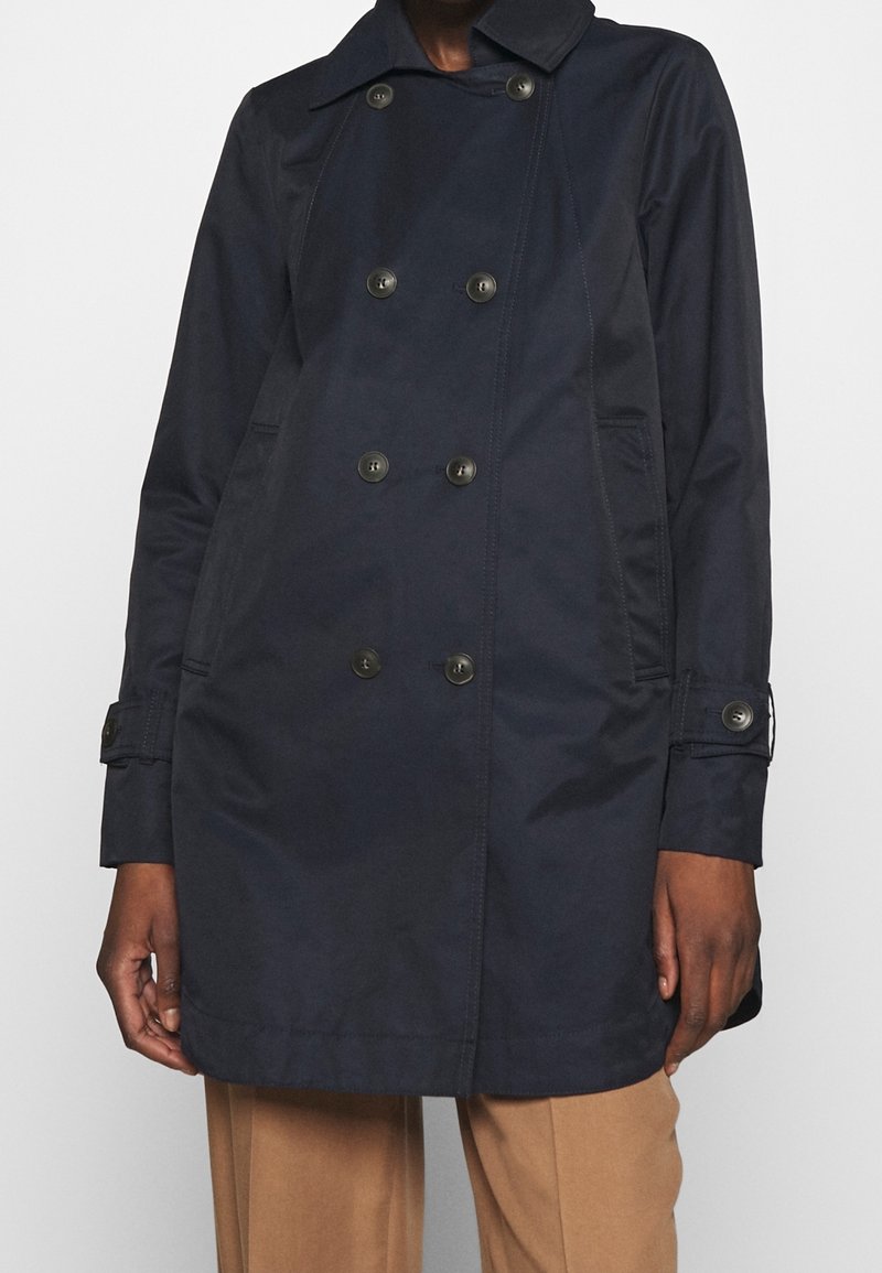 Abrigo azul marino de doble botonadura con detalles en botones, mangas largas y bolsillos laterales, llevado sobre pantalones beige.