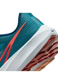 Tyrkysové běžecké boty s červenými detaily, texturovaný svršek, prodyšná síťovina a bílá mezipodešev s vzorovanou hnědou a oranžovou podešví.