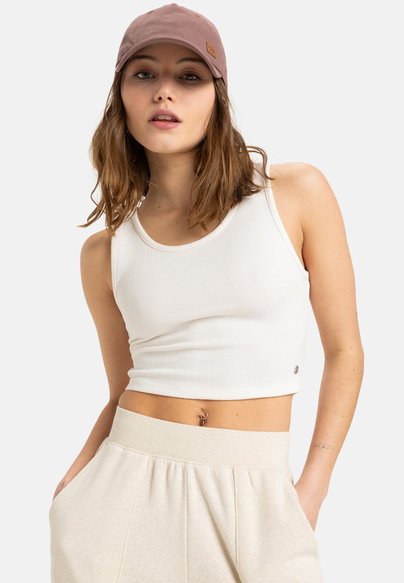 White cropped tank top with a textured finish, paired with light beige high-waisted sweatpants. A mauve cap complements the outfit.