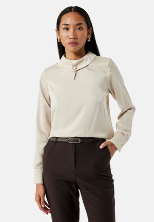 Crème blouse met een ronde kraag en een enkele parelknoop; lange mouwen met knoopmanchetten, gecombineerd met donkerbruine broeken.