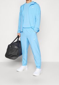 Lichtblauwe sportoutfit bestaande uit een zip-up jas en bijpassende joggers, gemaakt van een gladde stof. Bevat een zwarte sporttas en witte sneakers.