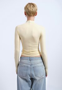 Pitkähihainen, vaaleankeltainen paita, jossa on napakka muotoilu, sileä tekstuuri, korkea kaula-aukko ja rypytetty yksityiskohta vyötäröllä, yhdistettynä korkeavyötäröisiin farkkuihin.