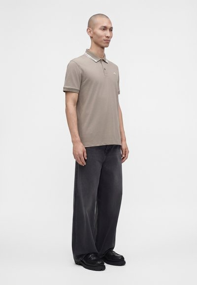 Homme debout portant un polo beige, un jean noir large et des chaussures noires sur un fond blanc uni.