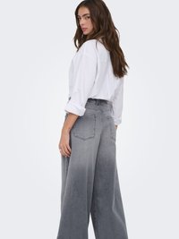 Mujer con cabello largo y castaño que lleva una camisa blanca abotonada y vaqueros grises de talle alto y pierna ancha, mirando por encima del hombro.