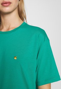 T-shirt en coton vert avec un col rond, des manches courtes et un détail logo doré sur la poitrine gauche. Texturé lisse et coupe décontractée.