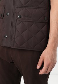 Καπιτονέ καφέ γιλέκο με δύο τσέπες στο στήθος, κλείσιμο με κουμπί και ανάγλυφο λογότυπο. Φορεμένο πάνω από σκούρες καφέ παντελόνες, που αναδεικνύουν ματ υφή.