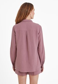 Woman with long brown hair wearing a mauve textured long-sleeve shirt and matching shorts, standing facing away against a white background.