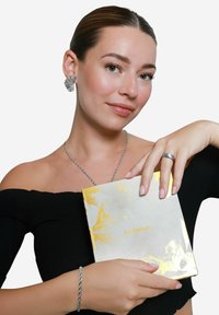Zilveren oorbellen in de vorm van bladeren, een zilveren ketting, een gevlochten zilveren armband en een boekomslag met gouden accenten, vastgehouden in een zwarte off-shoulder top.