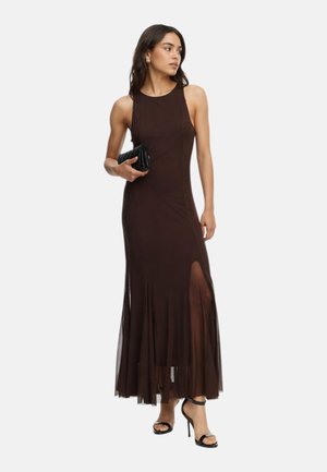 Donna in abito da sera marrone scuro senza maniche con spacco laterale che tiene una pochette nera, indossa sandali neri con tacco alto.