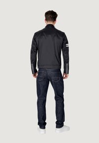 Sort nylonjakke med høj krave, med reflekterende striber på ærmerne, parret med mørke denimjeans og hvide sneakers.