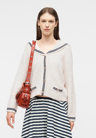 Kvinde iført en hvid kabelstrikket cardigan med navy kant, en stribet navy og hvid nederdel samt en rød lædertaske over skulderen.