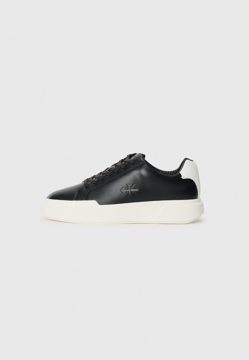 Adidași din piele neagră cu talpă din cauciuc alb. Prezintă vârf rotund, design cu șireturi și detalii cu logo pe lateral. Accentele din spate sunt albe.