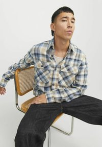 Karierter Flanellhemd in Blau, Beige und Grau mit zwei Brusttaschen, Knopfleiste und langen Ärmeln, kombiniert mit dunklen Jeans.