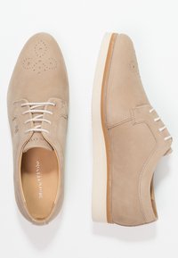 Chaussures en suede beige avec un bout arrondi, perforations décoratives sur le capuchon et lacets blancs. Présentent une semelle en caoutchouc de couleur claire.