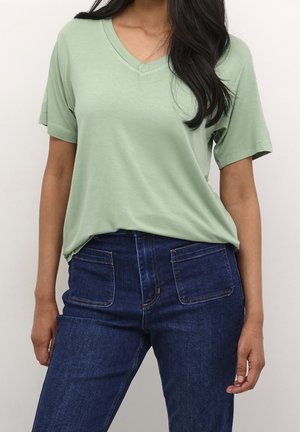 Femme portant un T-shirt vert clair à col en V et manches courtes, rentré dans un jean bleu foncé taille haute avec des poches plaquées devant.