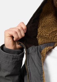 Grijze jas met een ritssluiting, voorzien van een bruine, fluffy voering en een geribbelde kraag. Een hand grijpt de ritssluiting, waardoor de textuur zichtbaar is.