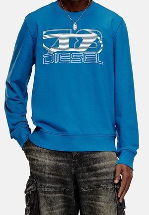 Sweat-shirt bleu avec logo Diesel blanc porté avec un jean cargo noir délavé, main dans la poche, et un collier argenté avec pendentif ovale.