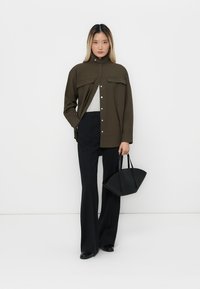 Olivovo zelená button-up bunda s dvoma náprsními vreckami, šedý top, čierne rozšírené nohavice a čierna štruktúrovaná kabelka. Hladká tkanina, moderný dizajn.