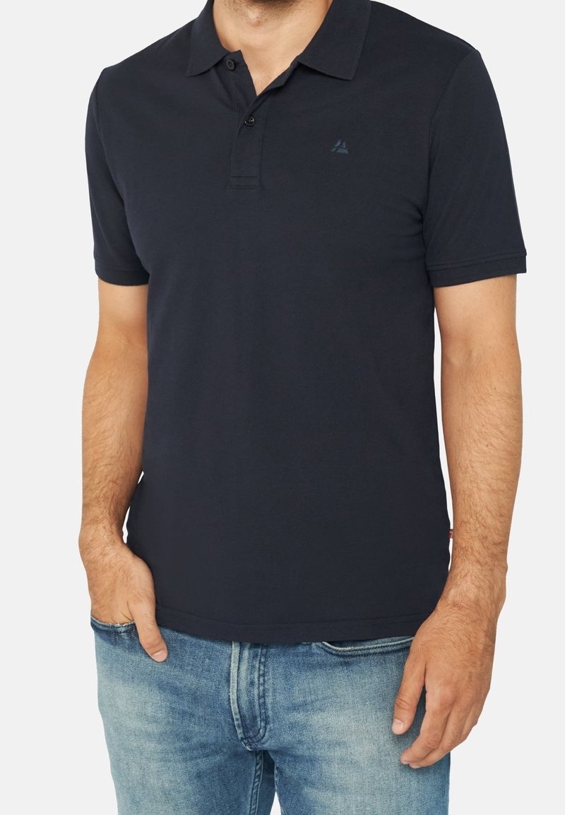 Mörkblå pikétröja i bomull; har en krage, treknappslås och en liten logotyp på vänster bröst. Strukturerad tyg.