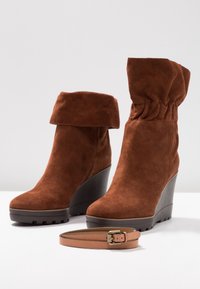 Bruna mockakängor med kilklack till vristerna, med en vikbar kant, rund tå och texturerad kilklack; tillsammans med ett tunt brunt bälte med ett guldspänne.