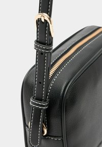 Zwarte leren handtas met een gouden ritssluiting, een verstelbare schouderband met witte stiksels en een gestructureerde afwerking.