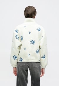 Fleecegenser i kremhvitt med blå blomsterbroderi, høy krage og lange ermer, med teksturert stoff og løs passform.