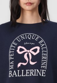 Jūras zila kapuce, kurai ir liels rozā lēdija priekšmets ar tekstu "MA PETITE UNIQUE BALLERINE" un "tici sev" baltā fontā.