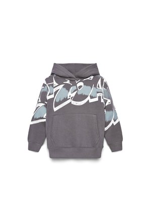 Giovane ragazzo in piedi che indossa una felpa con cappuccio grigia con grafica, pantaloni neri larghi e sneaker bianche e nere su sfondo grigio chiaro.