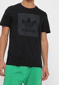 Svart kortärmad t-shirt i bomull med en matt svart Adidas-logotyp inuti en fyrkant med tandade kanter. Bärs med gröna shorts.