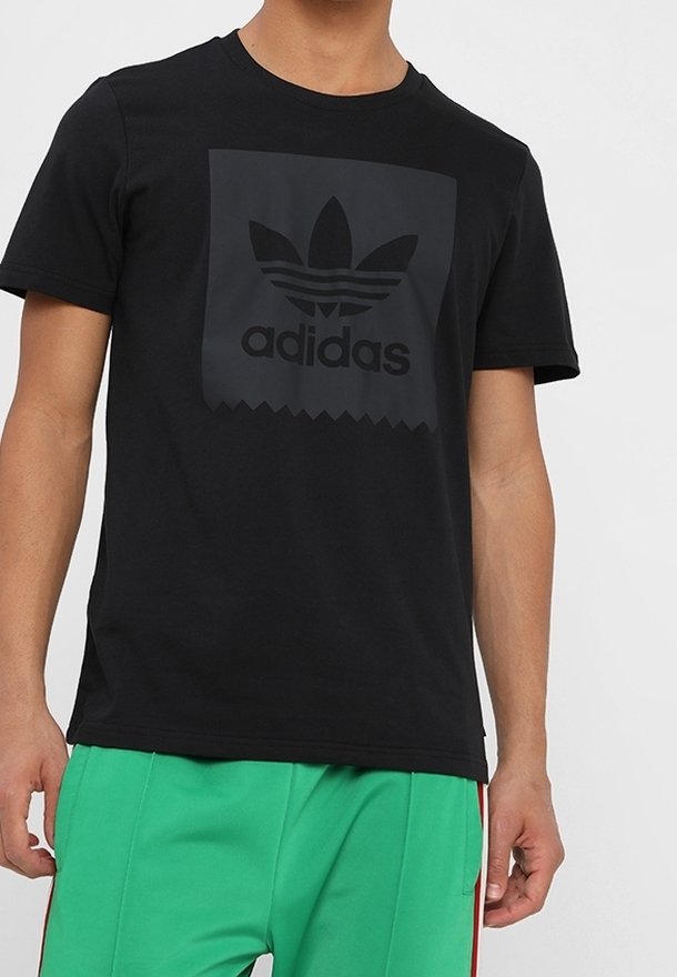 Svart kortärmad t-shirt i bomull med en matt svart Adidas-logotyp inuti en fyrkant med tandade kanter. Bärs med gröna shorts.