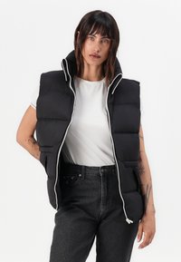 Svart polstret vest med høy krage, frontlukking med glidelås, og kontrasterende hvit kant. Båret over en hvit t-skjorte og svarte jeans.