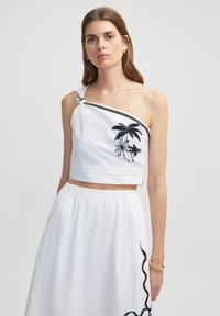 Witte cropped top met één schouder, voorzien van zwarte palmboom borduursels en een zwart touwaccent. Gecombineerd met een witte rok met elastische taille.