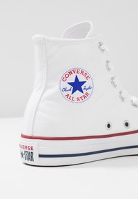 Bílé plátěné vysoké tenisky s kulatým modro-červeným logem "Converse All Star", bílými tkaničkami a červenobílým pruhovaným podešví.