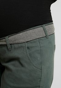 Gröna byxor med ett texturerat, svart och vitt herringbone-bälte. Har sidofickor och en slät yta. Fokus på midjebandsdesign.