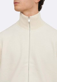 Cremfarbener Reißverschluss-Pullover mit hohem Kragen, der über einen strukturierten Strickhals und einen glatten Stoffkörper verfügt. Silberner Reißverschluss mit dezentem Glanz.