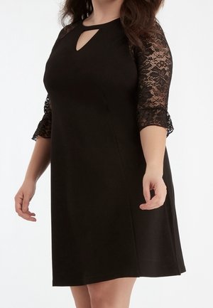 Frau trägt ein schwarzes knielanges Kleid mit Schlitz am Ausschnitt und dreiviertellangen Spitzenärmeln, posiert vor einem weißen Hintergrund.