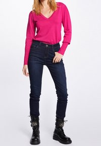 Fuchsia Strickpullover mit langen Ärmeln und V-Ausschnitt, kombiniert mit dunkelblauen, hochgeschnittenen Jeans; Modell trägt schwarze Schnürkampfstiefel mit metallischen Akzenten.