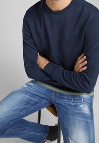 Mörkblå sweatshirt med rund hals. Bärs över en ljusgrön skjorta, ihop med slitna blåjeans. Minimalistisk design, avslappnad passform.