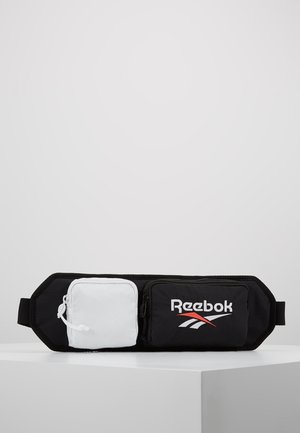 Sac banane Reebok noir et blanc avec deux compartiments zippés, un noir avec logo et un blanc uni, posé sur une surface blanche avec un fond gris.