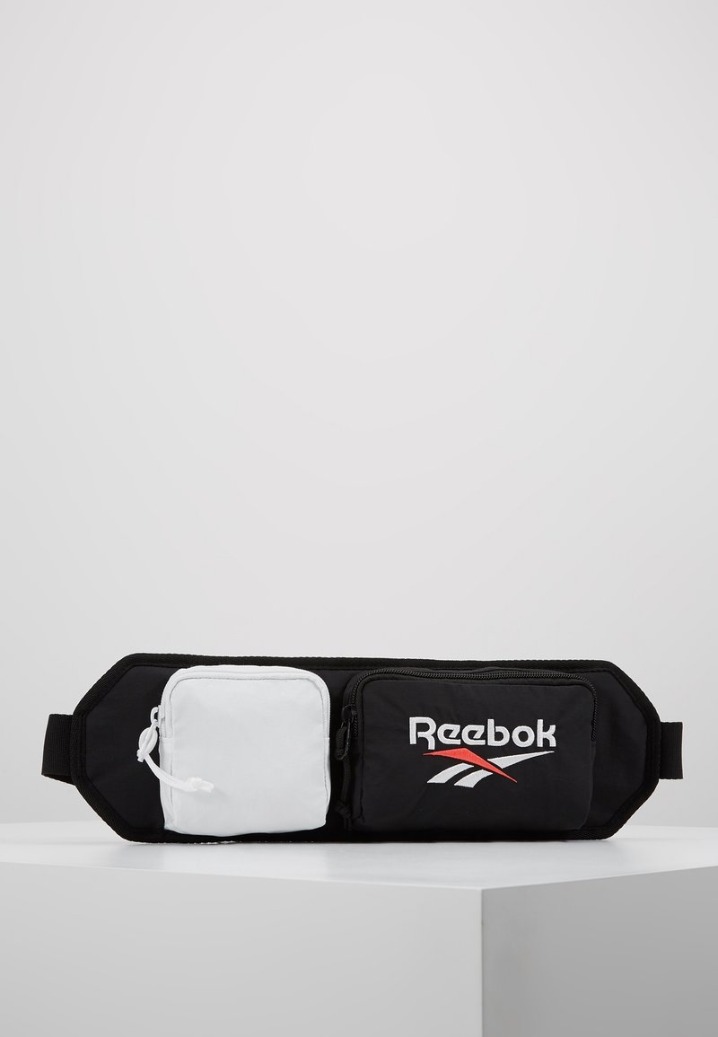Sac banane Reebok noir et blanc avec deux compartiments zippés, un noir avec logo et un blanc uni, posé sur une surface blanche avec un fond gris.