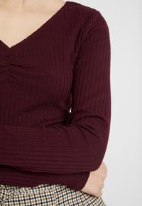 Osoba v proužkové vínové dlouhé tričko s řaseným výstřihem a kostkované sukni, se jednu ruku zkříženou přes pas.