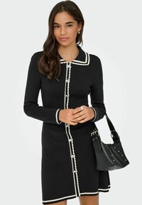 Crna pletena haljina s bijelim oštrim ovratnikom i manžetama, dugim rukavima, dugmadi sprijeda i lepršavim rubom. Crna torba s čavlima.