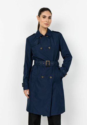 Femeie cu păr închis la culoare, purtând o haină lungă bleumarin cu curea și nasturi dubli, stând cu o mână în buzunar, pe un fundal alb.