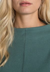 Zielony prążkowany top z okrągłym dekoltem i widocznymi detalami szwów, w połączeniu z długimi falowanymi blond włosami i neutralnymi ustami.